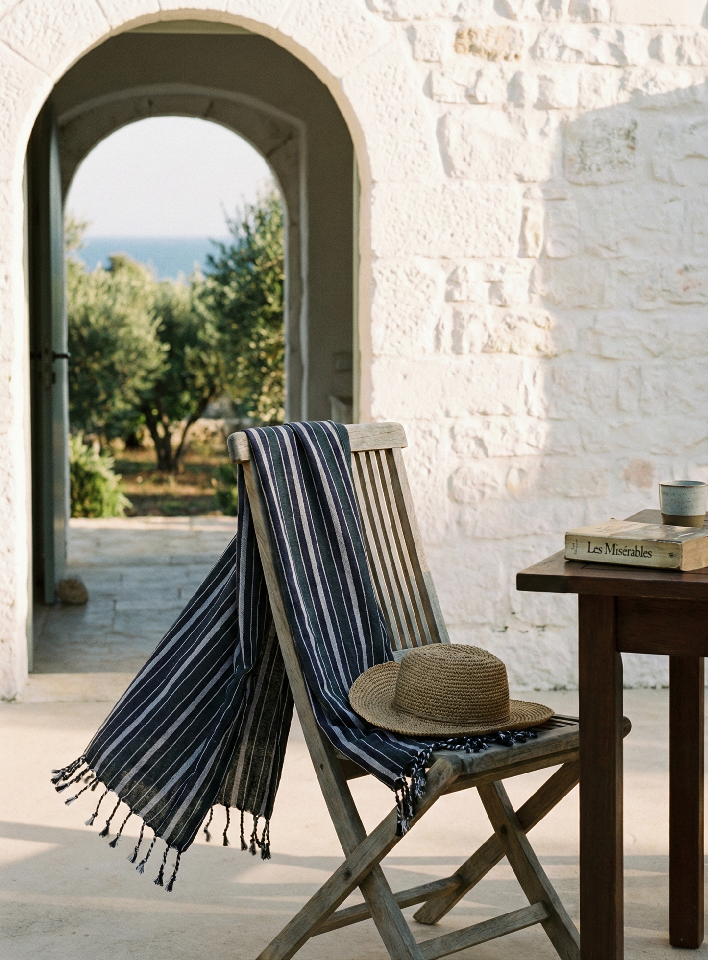 Chair with a striped towel and straw hat on a patio with a table and view of trees.